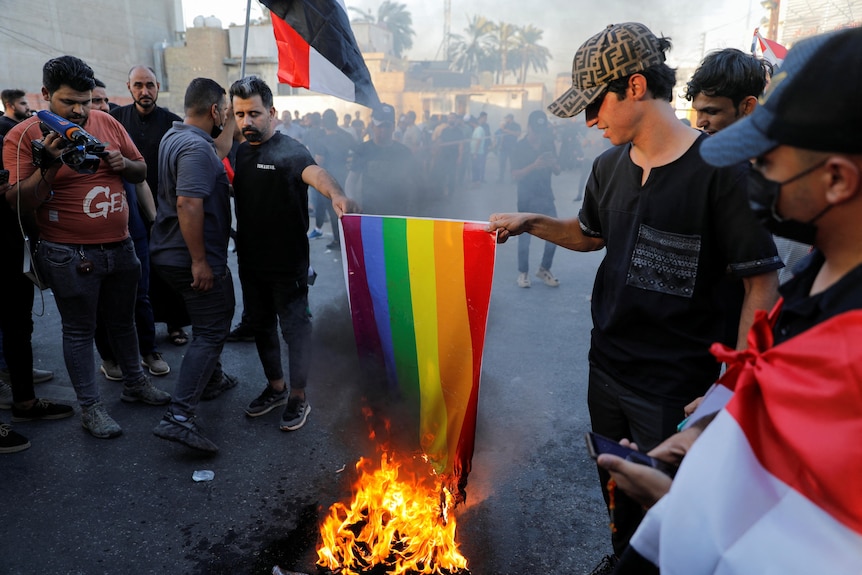 a rainbow flag being lowered into a fire on the floor by two people among a crowd