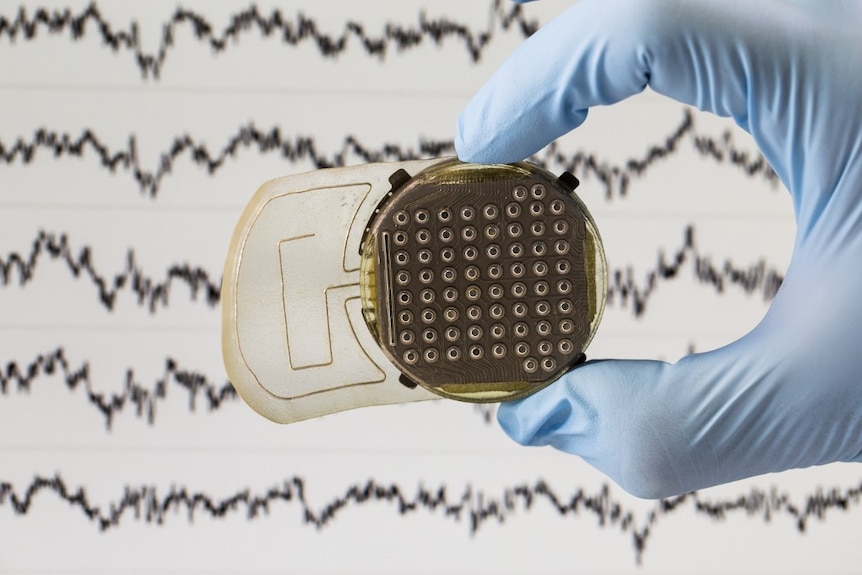 A brain implant is held up in front of a scan of brainwaves
