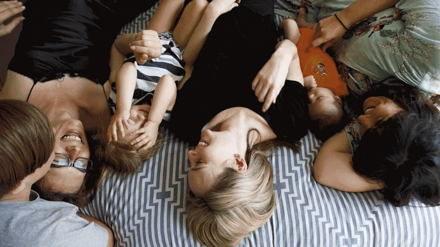 Quote: "It works because it's not share housing in a traditional sense." Pic: Three women and kids lie on bed smiling.