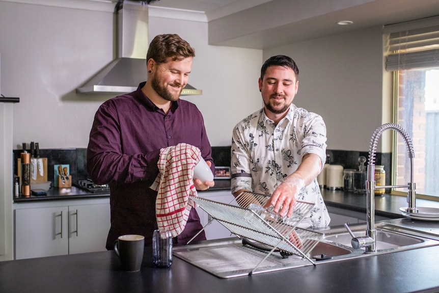 Two men wearing button-down shirts wash dishes in a household kitchen.