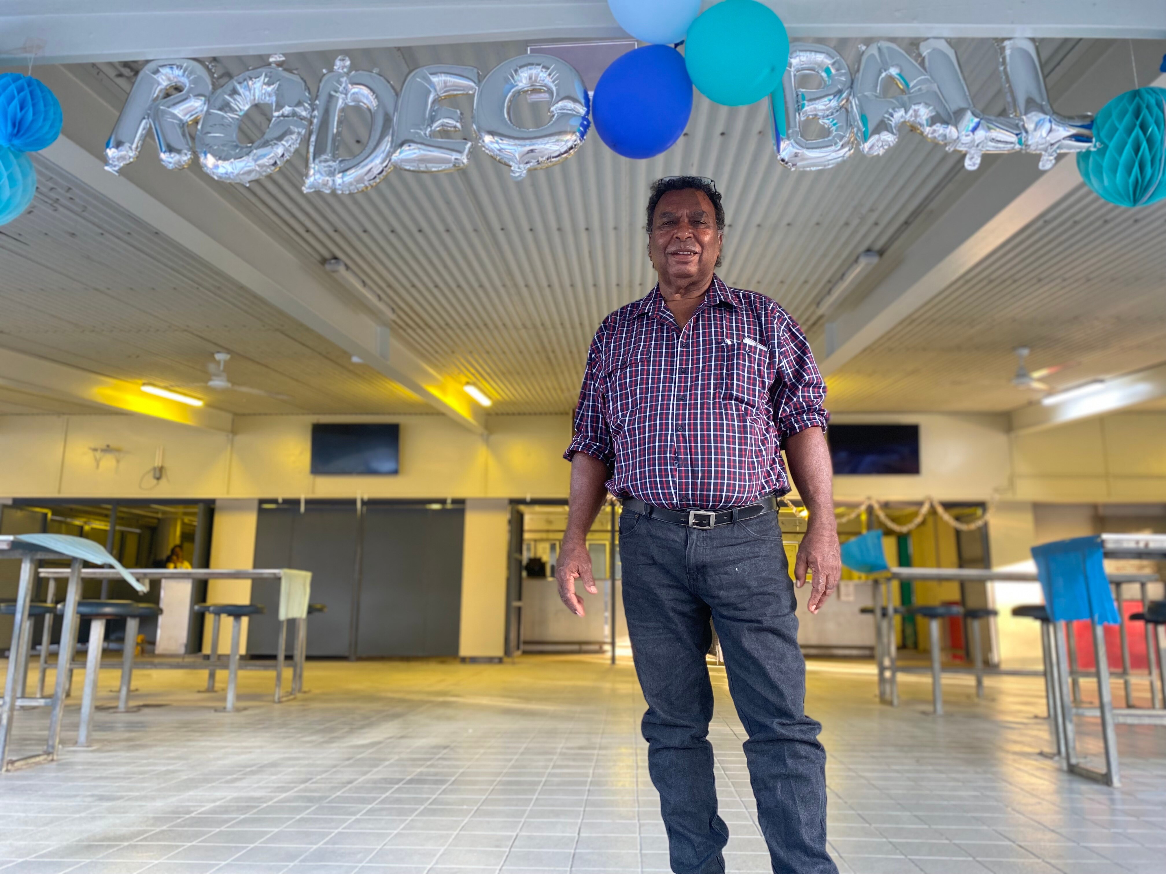An Aboriginal man dressed in jeans and a plaid shirt stands under a string of balloons reading 'Rodeo Ball'.