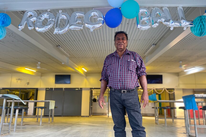 An Aboriginal man dressed in jeans and a plaid shirt stands under a string of balloons reading 'Rodeo Ball'.