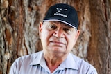 A head shot of an older Indigenous man, wearing a blue cap, sky blue t-shirt, stands against a tree bark, looks serious.