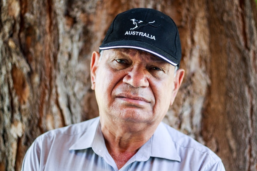 A head shot of an older Indigenous man, wearing a blue cap, sky blue t-shirt, stands against a tree bark, looks serious.