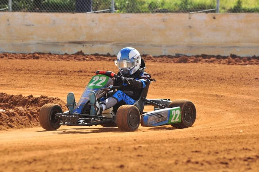 a young boy rides in a blue and green go-kart, he wears a black and blue race suit and a blue and white helmet, he races on dirt