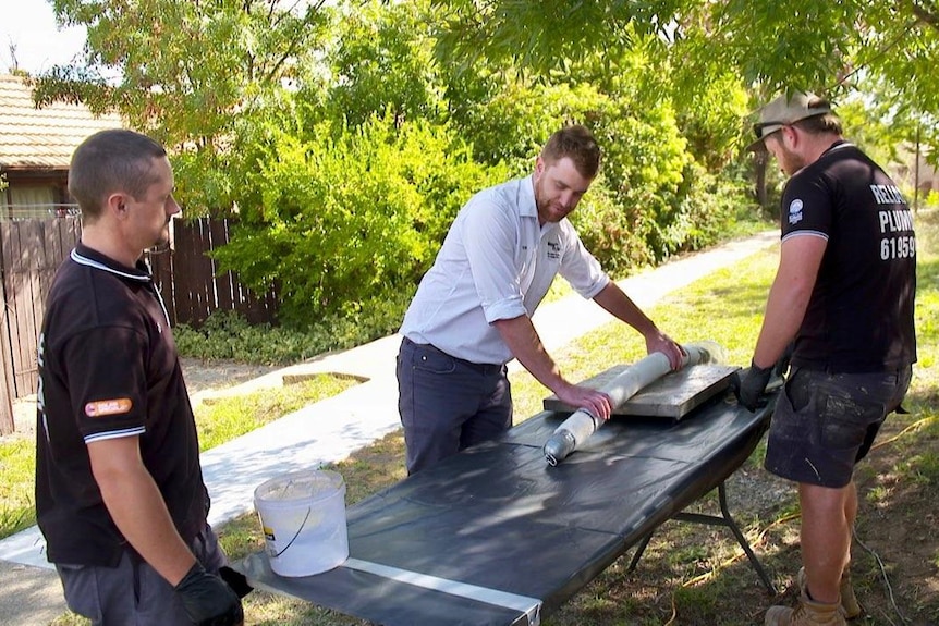 Three men use a workbench to bend a piece of plumbing pipe.