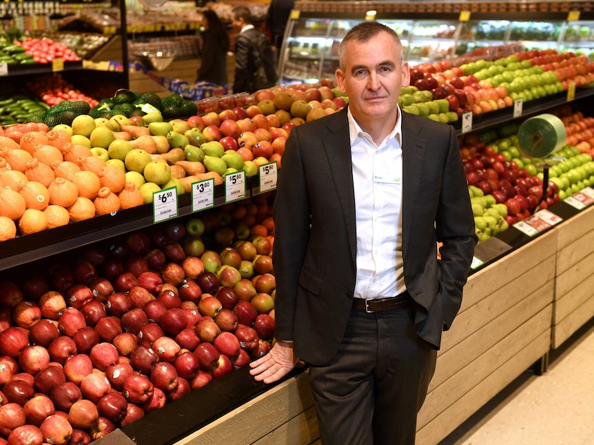 A man wearing a suit stands besides a fresh fruit stand in a supermarket.