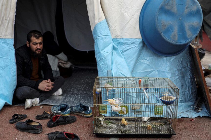 A man sits at a door of a tent looking at a bird in a cage. 