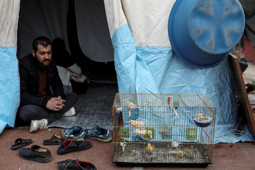 A man sits at a door of a tent looking at a bird in a cage. 