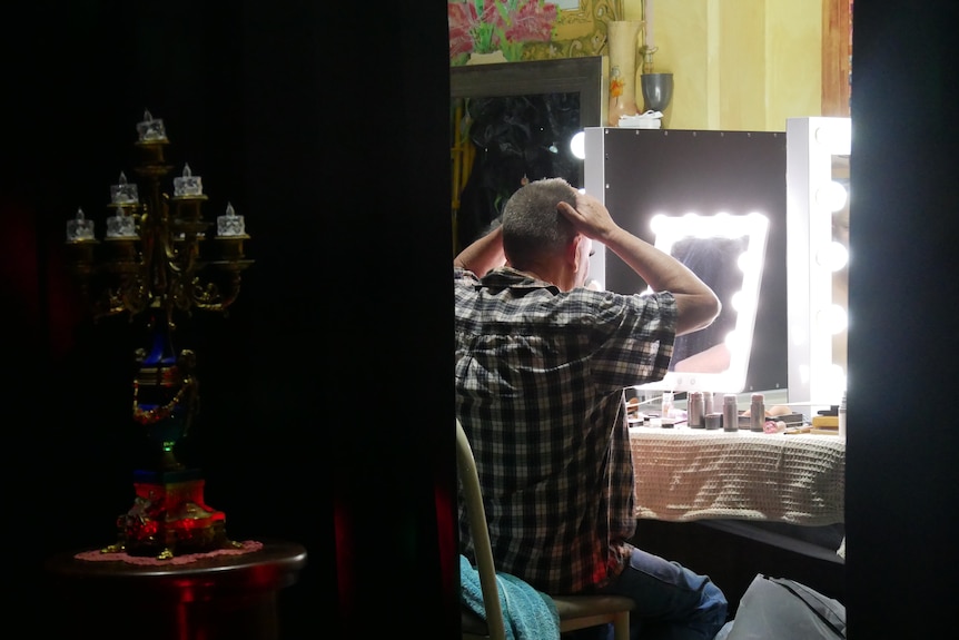 Behind a velvet curtain and candelabra, a man looks in a mirror and adjusts his hair. 
