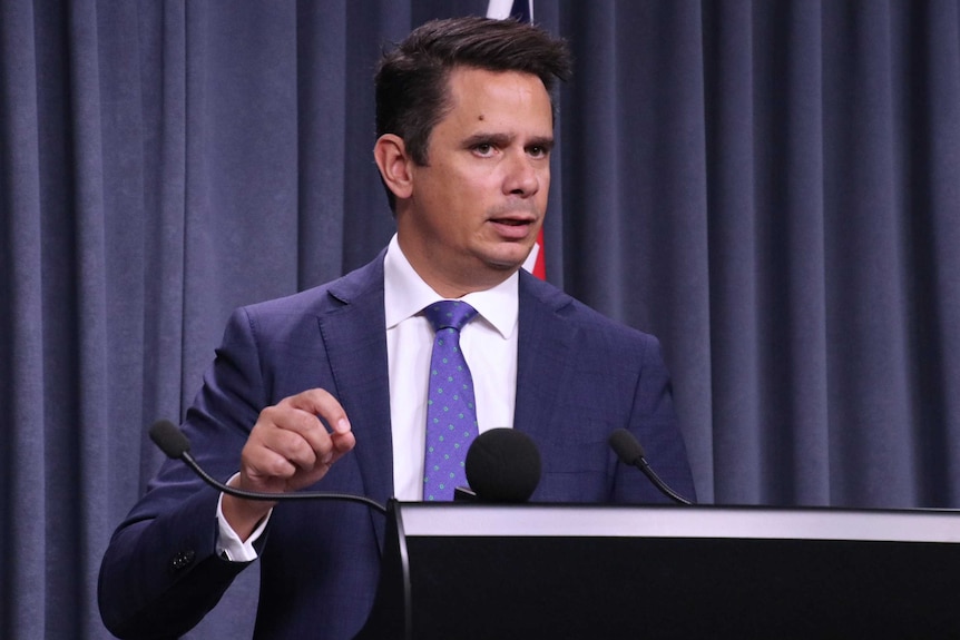 A mid-shot of WA Treasurer Ben Wyatt in a suit and tie standing at a dais talking into three microphones indoors.