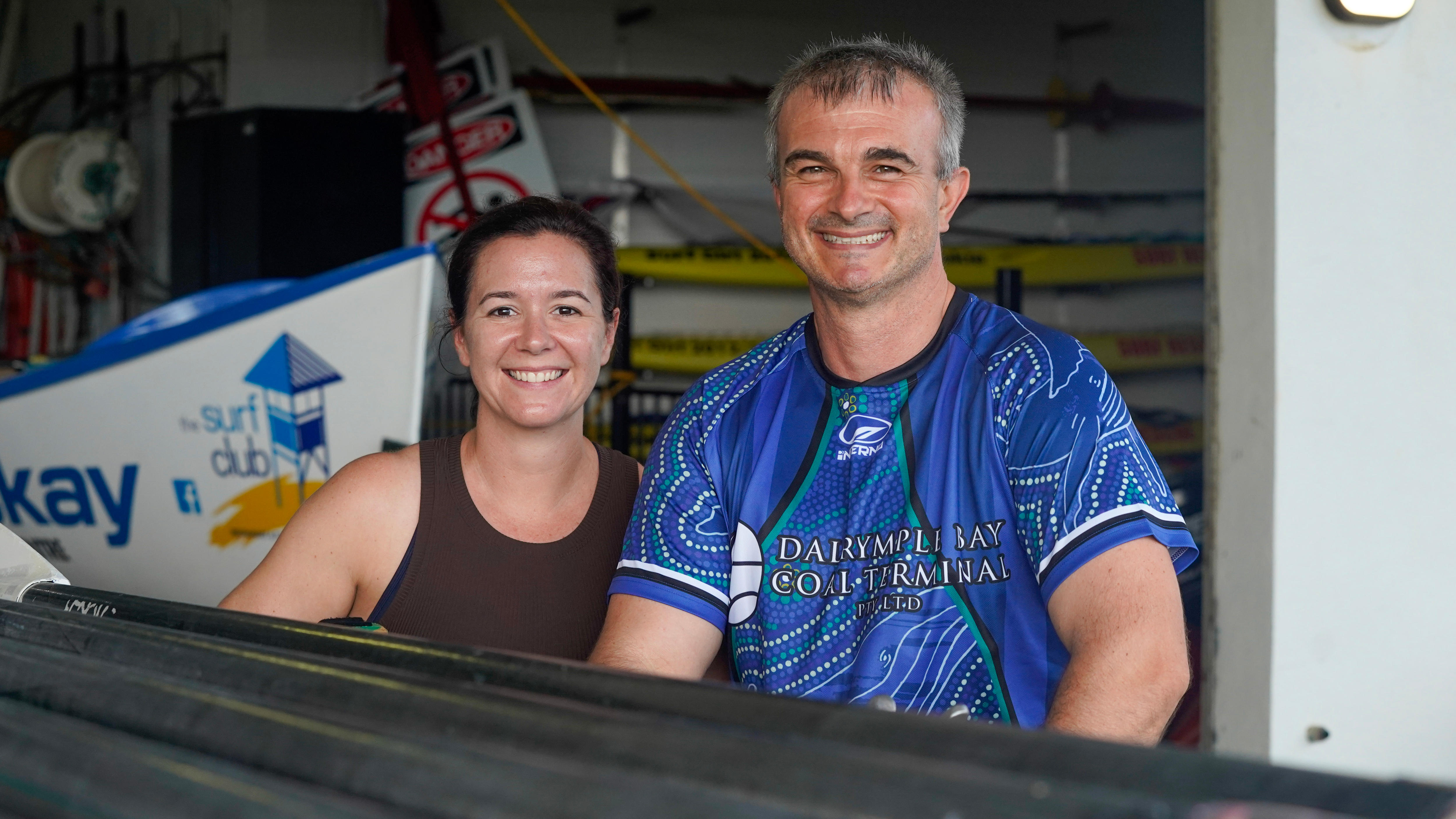 Una mujer y un hombre sonrientes se apoyan en una pila de remos negros sobre un estante en un cobertizo para botes.