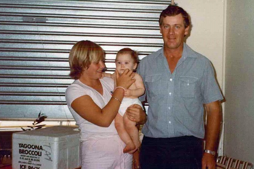 A 1980s photograph of a man standing with his two daughters. one is a baby being held by her sister