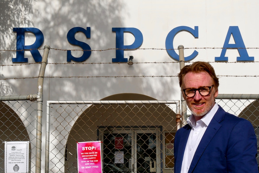 A man with red hair wearing a blue suit and glasses in front of the Broken Hill RSPCA clinic. 