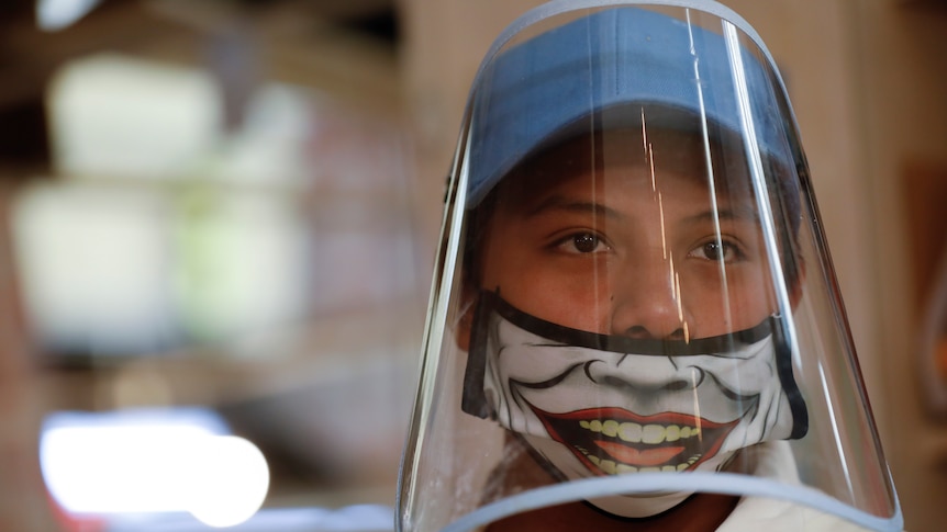 A boy wears a plastic face shield over a Joker face mask.