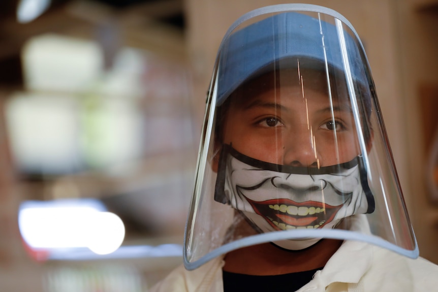 A boy wears a plastic face shield over a Joker face mask.