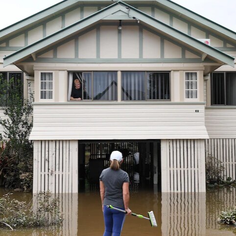A woman looks out of her first story window as flood water laps at hair stair case, her whole yard covered in brown water.