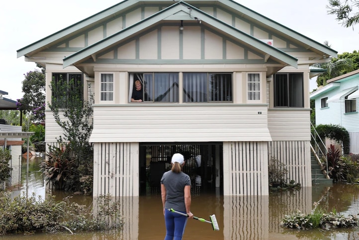 A woman looks out of her first story window as flood water laps at hair stair case, her whole yard covered in brown water.