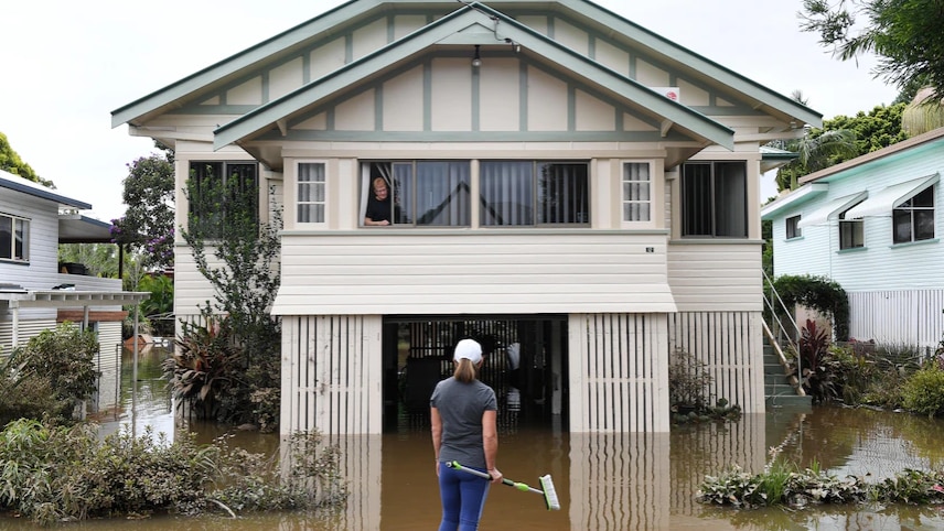 A woman looks out of her first-storey window as flood water laps at her staircase, her whole yard covered in brown water.