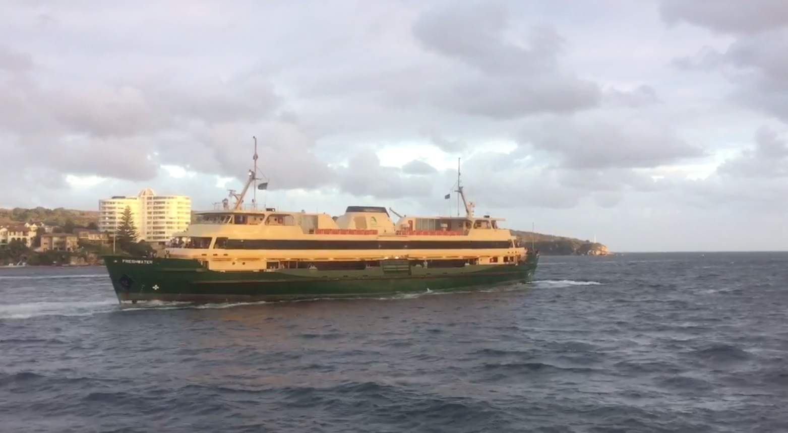 A green and cream ferry on the waters, high-rise buildings can be seen in the distance.