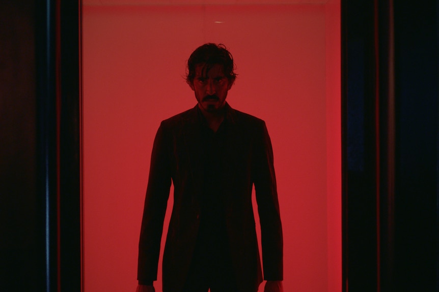 A film still of Dev Patel. He is lit by red light behind him, and is wearing a suit and a determined expression.