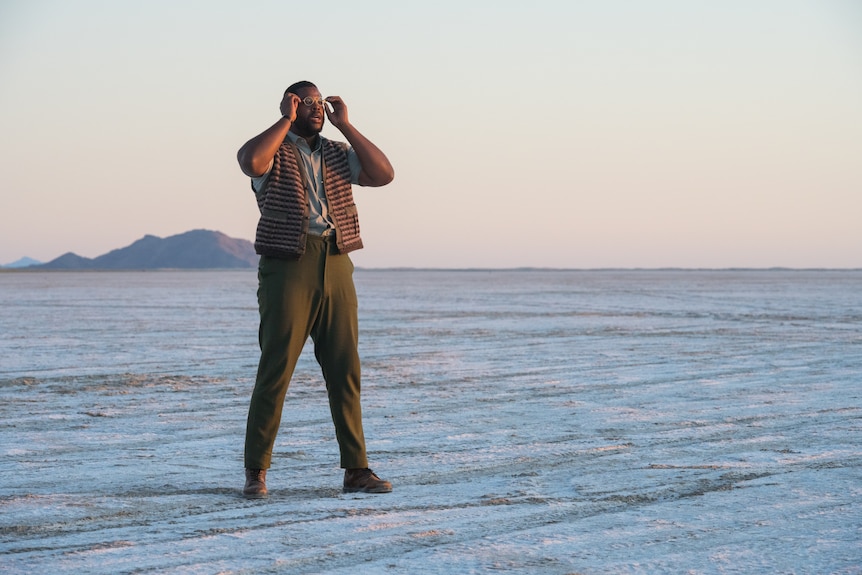 A tall Black man wearing trousers and a vest holds his hands up to his glasses as he stands in a grey desert