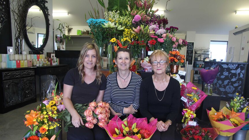 Three women sit among colourful flowers in a shop