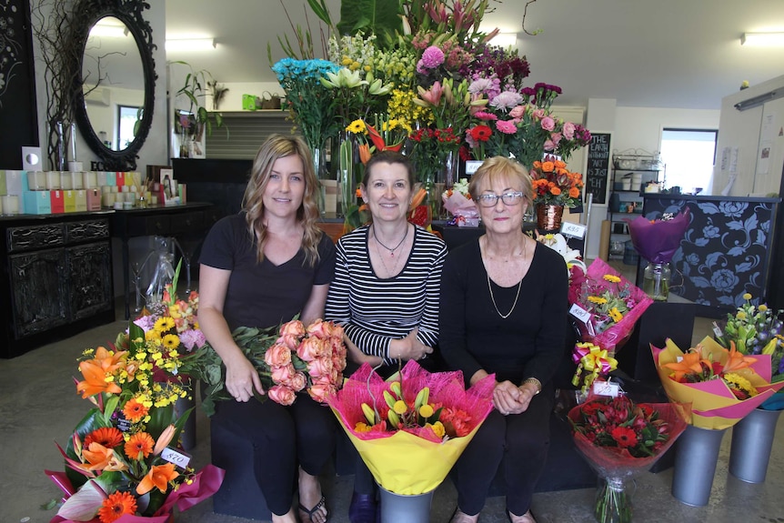 Three women sit among colourful flowers in a shop