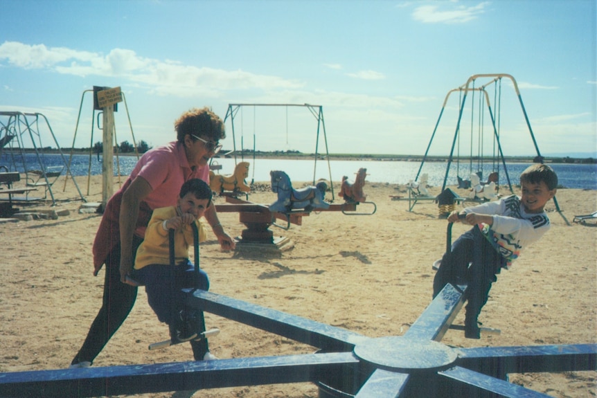An old coloured film photo of a lady wearing a pink polo pushing her grandkids on a see saw at the beach playground