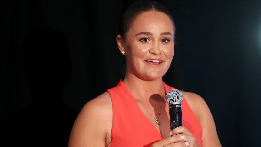 A smiling Ash Barty stands on stage holding a microphone during a presentation.