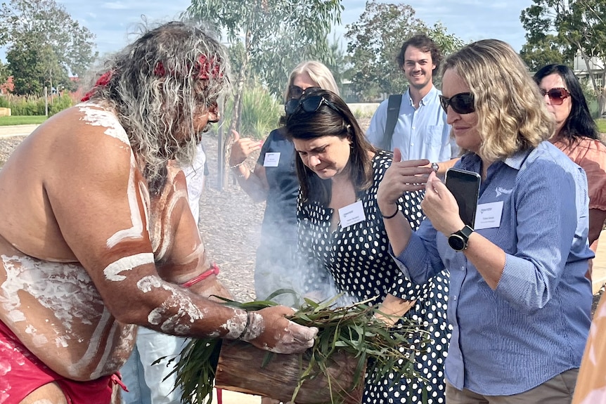 An Indigenous man holds a coolaman with smoking Eucalyptus leaves up to a woman.