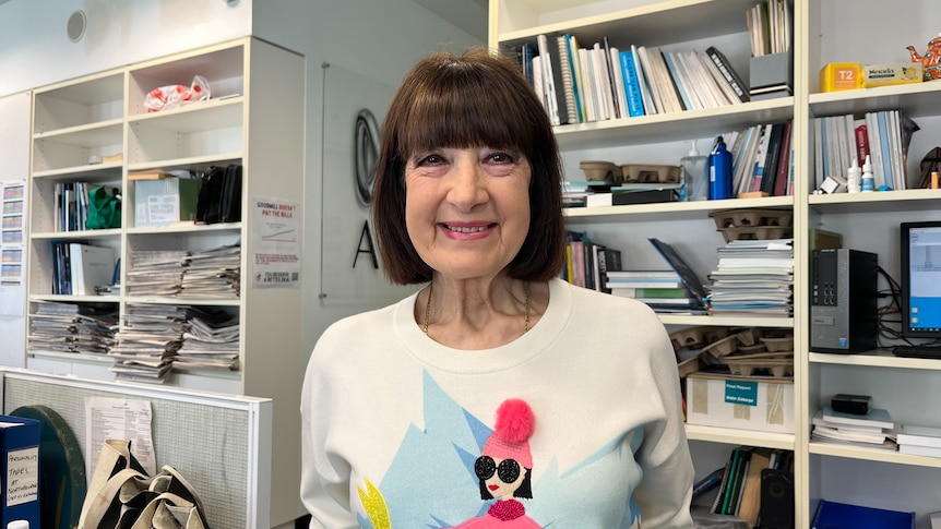middle-aged woman stands smiles at the camera, she has a short bob cut and bangs and wears a white jumper with a funky print