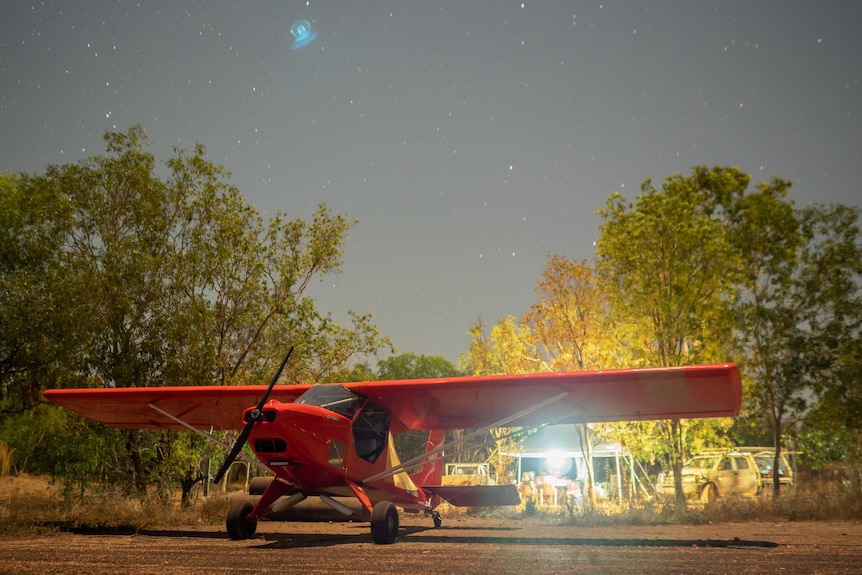 A small red plans sits on dirt under a starry night in front of some large trees.