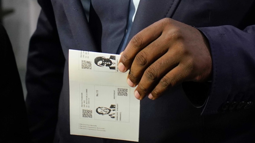 A close-up on a man's hand holding a ballot paper with two men's faces depicted.