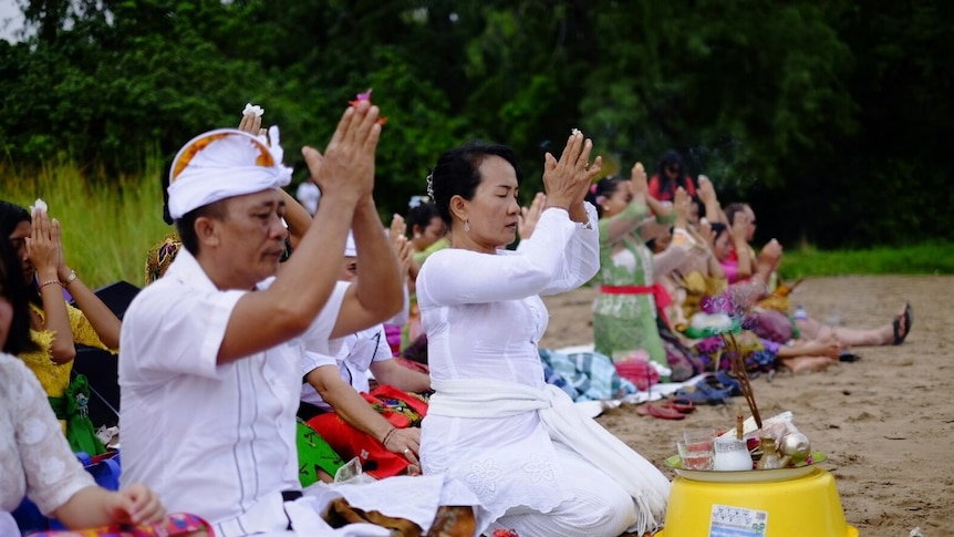 A line of Balinese people sitting in the sand dressed in traditional costumes as they pray.