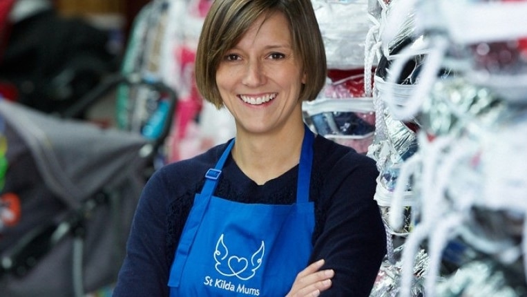 A woman wearing a blue St Kilda Mums apron stands in front of piles of packaged clothing and donated goods