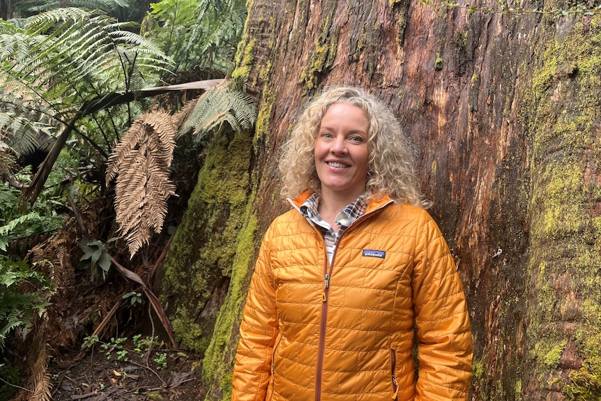 A blonde woman wearing a yellow jacket standing in front of some ferns and a large tree