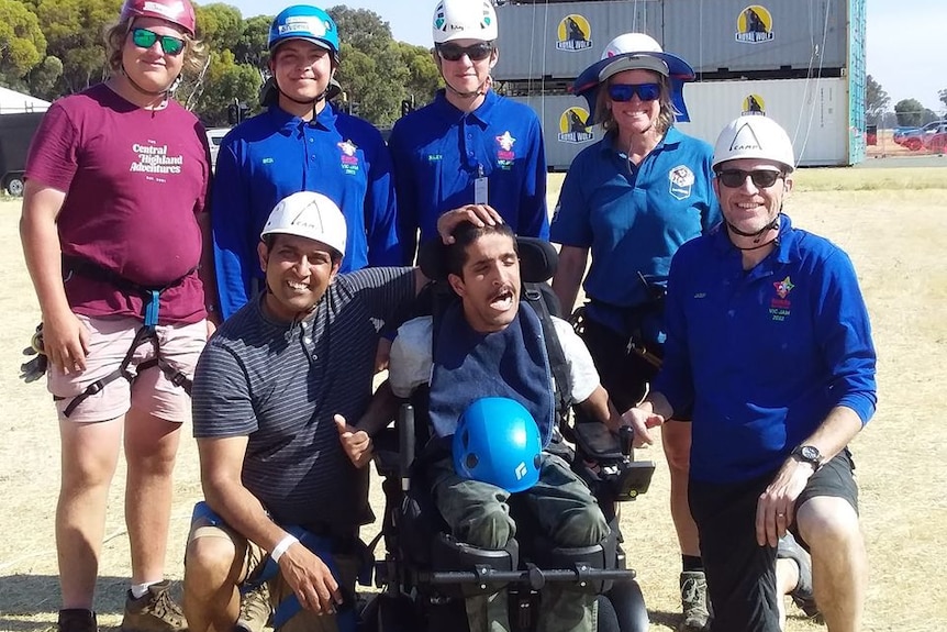 Four people with blue t shirts and helmets, and two others are standing and kneeling around a man in a wheelchair.