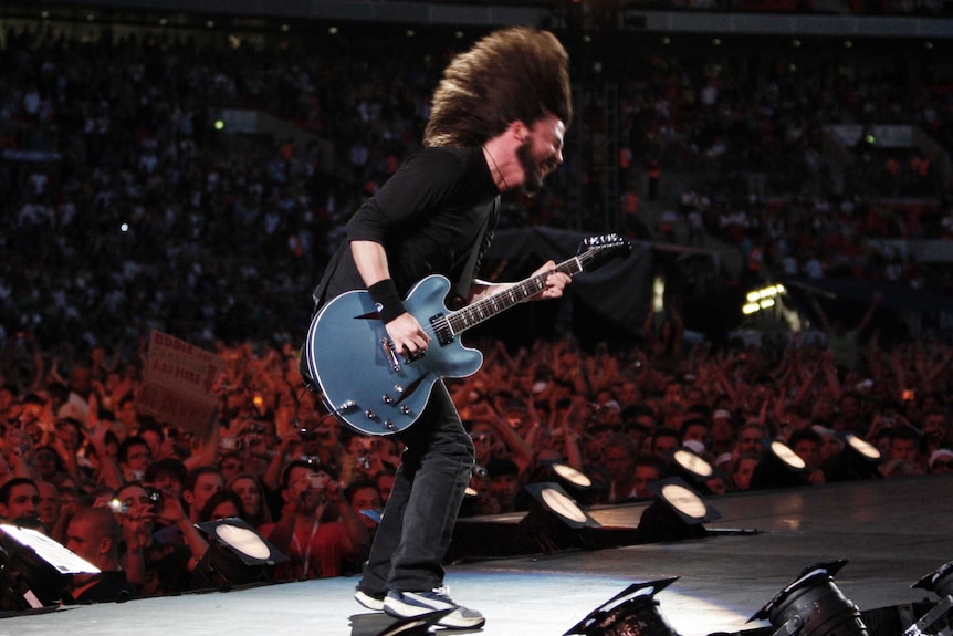Dave Grohl, mid-head bang and with mouth open, playing the guitar on stage with a large stadium crowd in the background