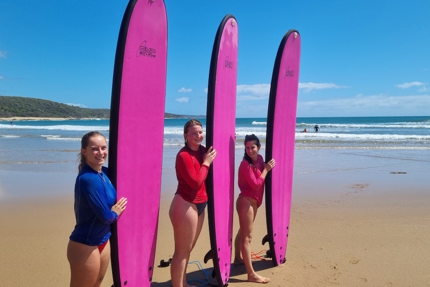 Four young women holding bright pink surfboards on the beach at Agnes Water.