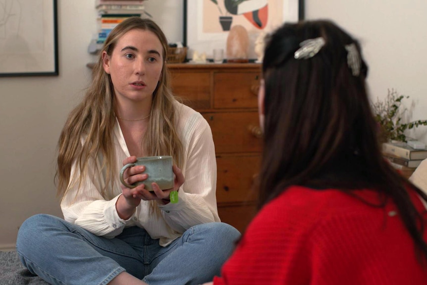 Lauren holds a mug while sitting on a bed talking with a friend.