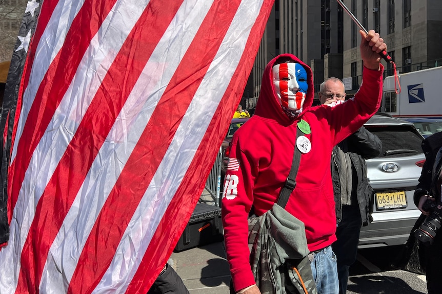 A man in a red hoodie wearing an American flag mask waves an American flag