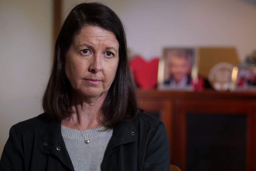 A concerned looking woman sits in her home with photographs of her mother on a sideboard behind her.