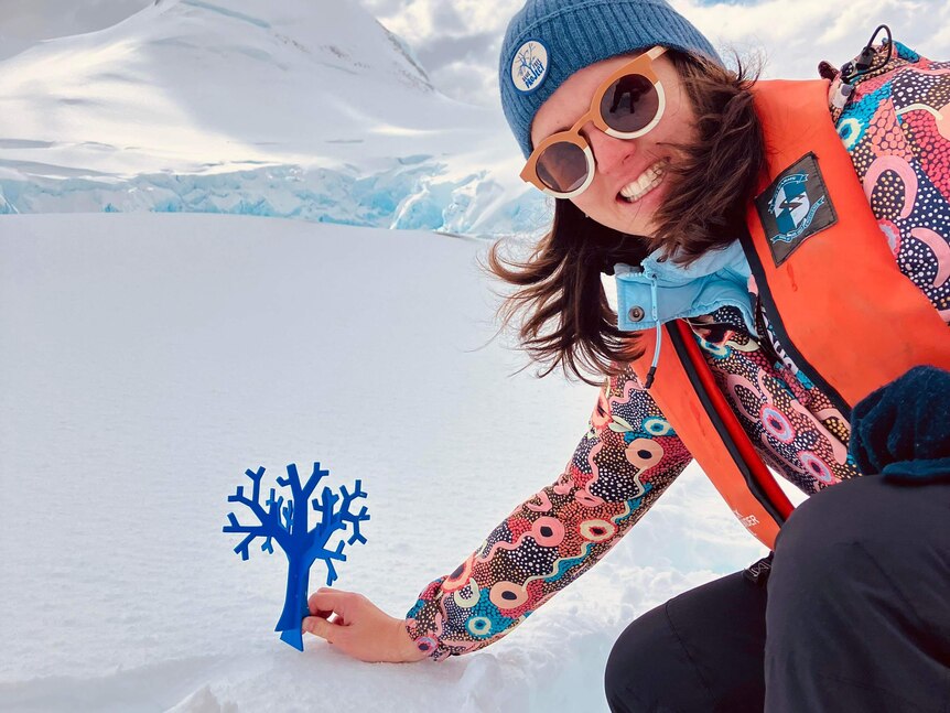A woman holds a small blue wooden tree on the snow in Antarctica.