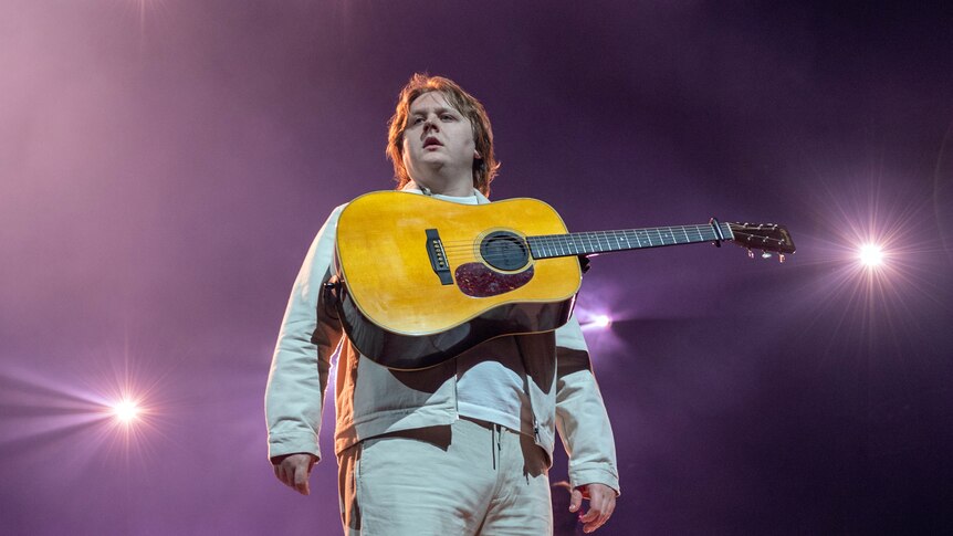 Lewis Capaldi stands on stage in a white shirt, pants and jacket and an acoustic guitar strapped high on his chest.