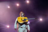 Lewis Capaldi stands on stage in a white shirt, pants and jacket and an acoustic guitar strapped high on his chest.