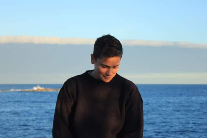Kaya Wilson, with short black hair and black jumper, looks down smiling with a backdrop of the blue ocean.