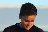 Close up shot of Kaya Wilson, with short dark hair and in black jumper, looking down and smiling slightly. The ocean is behind.