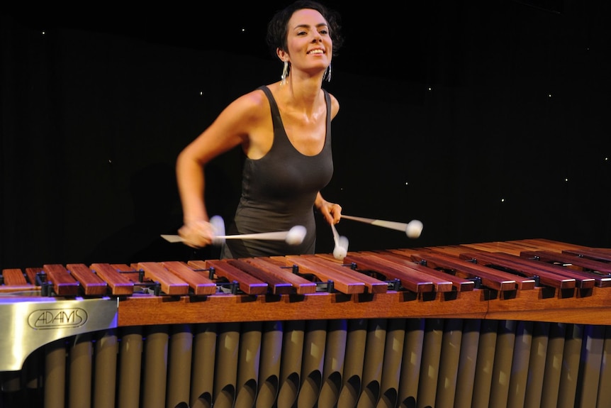 Claire Edwardes plays a marimba, holding four mallets and wearing a black singlet top.