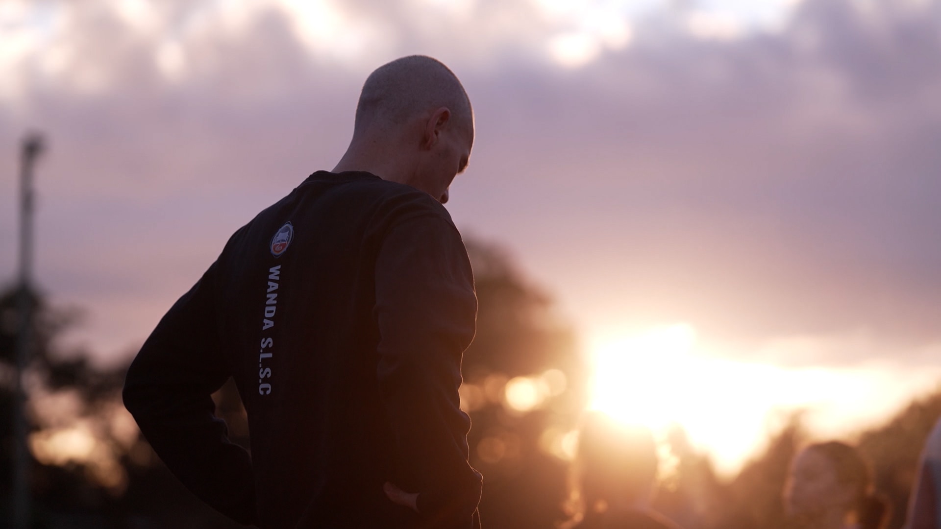 A man is seen from behind wearing a long-sleeved black tshirt and standing with his hands on his hips as the sun rises.